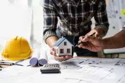 Architekci omawiają projekt domu, planując, jak uzyskać pozwolenie na budowę. Na stole makieta domu, plany, kask ochronny i kalkulator.