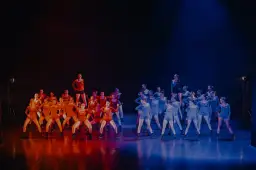 Grupa tancerzy na scenie, podzielona na dwie grupy: jedna w pomarańczowych strojach, druga w niebieskich. Nauka tańca nowoczesnego w pełnym blasku.