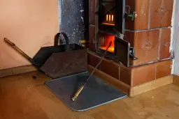 Piec kaflowy w ogniu, obok pogrzebacz i popielnica. Rozważasz rozbiórkę pieca kaflowego? Dofinansowanie może pomóc!