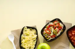 Dwa pojemniki z jedzeniem: makaron farfalle i sałatka z groszku, kapusty i papryki. Obok jabłko i łyżka.
