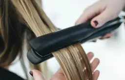 Dłoń trzyma prostownicę, wygładzając blond włosy. Nie wiadomo, ile stopni ma prostownica, ale efekt jest widoczny.