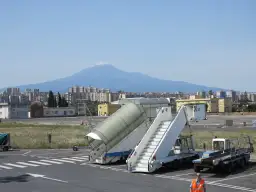 Port lotniczy Katania Fontanarossa z widokiem na Etnę. Schody lotnicze i pojazdy obsługi naziemnej na tle miasta i wulkanu.