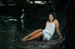 Kobieca sesja w wodzie. Kobieta w białej, mokrej sukience siedzi na błotnistym brzegu strumienia, otoczona korzeniami drzew.