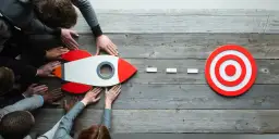 Rakieta i tarcza na drewnianym tle, otoczone dłońmi.