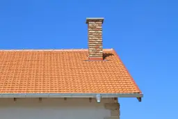 Ceglany komin na dachu z pomarańczowej dachówki. Zastanawiasz się, ile kosztuje budowa komina?