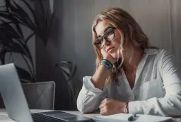 Kobieta w okularach opiera głowę na dłoni, patrząc zamyślona na laptopa.