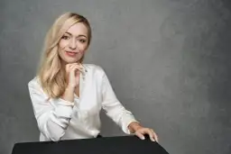 Blondynka w białej satynowej koszuli podczas sesji modowej, z dłonią pod brodą, uśmiecha się do obiektywu.