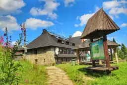 Kamienny schronisko na Turbaczu z dzieckiem w tle, otoczone zielenią i kwitnącymi kwiatami.
