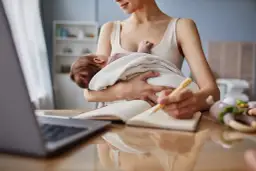 Kobieta trzyma dziecko i pisze w notatniku przy laptopie.
