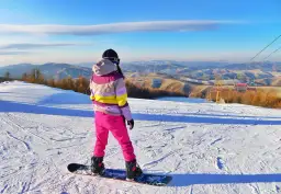 Snowboardzistka w różowych spodniach i kolorowej kurtce na tle górskiego krajobrazu.