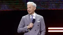 Mężczyzna w prążkowanym garniturze, prowadzący "Milionerzy", z mikrofonem w dłoni.