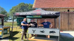 Trzech mężczyzn pod namiotem Koła Łowieckiego "Leśnik". Czy myśliwy może odsprzedać tuszę? Na grillu widnieją symbole zwierzyny.