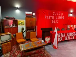 Scena imprezy w stylu PRL: meble, radio, telewizor, akordeon, czerwony transparent z napisem.