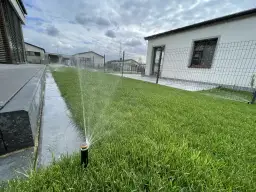 Zraszacz wynurzalny podlewa zielony trawnik obok domu.