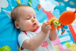 Niemowlę leżące na kolorowej macie bawi się zabawką.