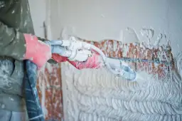 Ręce w czerwonych rękawicach nakładają tynk na ceglaną ścianę za pomocą agregatu.