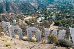 Widok zza gigantycznych liter znaku Hollywood. W oddali widać wzgórza z domami i drogami.