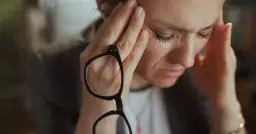 Kobieta z dłońmi na skroniach, trzymająca okulary, z wyrazem zmartwienia na twarzy.
