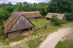 Drewniane chaty ze słomianymi dachami w skansenie, otoczone zielenią.