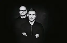 Dwóch mężczyzn w czarnych bluzach na ciemnym tle. Jeden z nich ma okulary, drugi irokeza. Wyglądają jakby właśnie wylądowali po locie dzika.