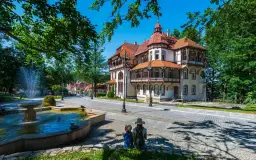 Dwie osoby siedzą przy fontannie, podziwiając architekturę luksusowego sanatorium w otoczeniu zieleni.