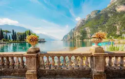 Kamienna balustrada z wazonami kwiatów, widok na turkusowe jezioro Garda i góry. Statek płynie po wodzie.