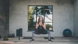 Kobieta wykonuje przysiad sumo. Widoczne hantle, piłka gimnastyczna i maty. Trening HIIT efekty widać po jej sylwetce.