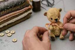 Mały, ręcznie robiony miś z materiału, ozdabiany wstążką, obok stosu tkanin i akcesoriów.