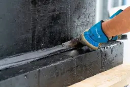 Rękawica z narzędziem nakłada czarną masę izolacyjną, chroniąc fundamenty przed wilgocią.