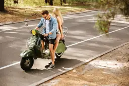 Młoda para na zielonym skuterze, ciesząca się przejażdżką po leśnej drodze. Kto może jeździć skuterem? Ci, którzy szukają wolności i przygody!