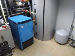 Niebieski, mały kocioł co marki Buderus obok dużego zasobnika i zbiornika wyrównawczego.