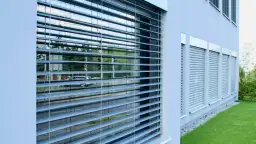 Nowoczesny budynek z jasną elewacją i oknami osłoniętymi przez rolety zewnętrzne ręczne. W odbiciu widać ulicę i samochód.