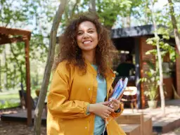 Uśmiechnięta dziewczyna o śniadej cerze, z kręconymi włosami, w żółtej kurtce, trzyma książki i słuchawki.