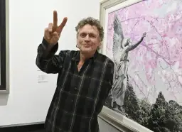 Rick Allen, perkusista Def Leppard, pozuje z uniesionym znakiem pokoju obok obrazu z aniołem i kwitnącymi drzewami.