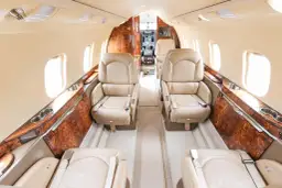 Luksusowe wnętrze samolotu czarterowego: wygodne, skórzane fotele w jasnym kolorze, eleganckie drewniane wykończenia i okna.