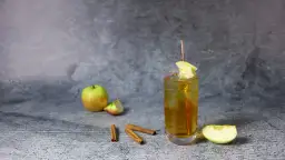 Szklanka z drinkiem, jabłka, laska cynamonu na szarym tle.