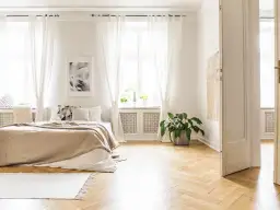 Sypialnia z łóżkiem w kolorze cappuccino, idealnie komponującym się z jasnymi zasłonami, drewnianą podłogą i zieloną rośliną.