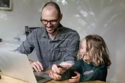 Ojciec z córką przy laptopie, pokazujący jej coś na telefonie. Usługa zdalna.
