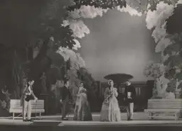 Scena z przedstawienia teatralnego, gdzie aktorzy w strojach z epoki odgrywają scenę w ogrodzie. Reżyser teatralny Szyfman zadbał o szczegóły scenografii.
