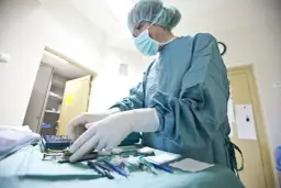 Chirurg w masce i rękawiczkach przygotowuje narzędzia chirurgiczne na stole operacyjnym.