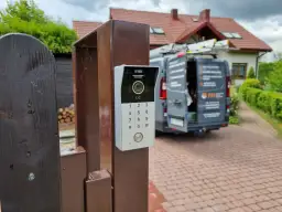 Nowy domofon na furtce. Zastanawiasz się, ile kosztuje montaż domofonu w domu jednorodzinnym? W tle widać dom i samochód firmy instalacyjnej.