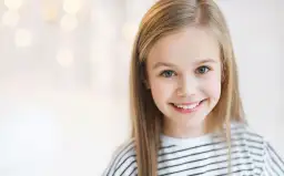 Uśmiechnięta dziewczynka o blond włosach, jakby inspirowana nowoczesnymi imionami dla dziewczyn, z rozmazanym tłem pełnym świateł.