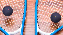 Dwie rakiety do squasha z czarnymi piłeczkami (jedna z czerwoną, druga z żółtą kropką) na drewnianym tle.