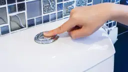 Dłoń naciska chromowany przycisk spłuczki WC na białej ceramice, w tle mozaika.