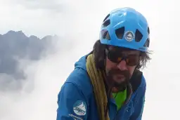 Mężczyzna w kasku i okularach przeciwsłonecznych, z liną wspinaczkową na szyi, na tle mglistych szczytów. Perć w Tatrach.