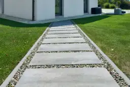 Ścieżka z prostokątnych płyt betonowych i kamieni ozdobnych obok trawnika i białego domu.
