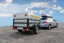 Srebrny samochód z boxem dachowym ciągnie przyczepę z widocznym ładunkiem.