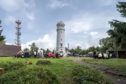 Ludzie odpoczywają przy wieży widokowej i maszcie telekomunikacyjnym. W oddali widać wielką sowę na tle chmur.