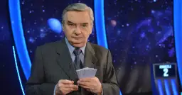 Starsza osoba w garniturze trzyma karty z napisem "1 z 10", gotowa do emisji.