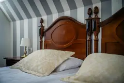 Drewniane łóżko z ozdobnymi wezgłowiami i poduszkami na tle tapety w paski.